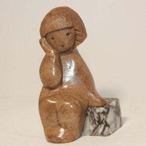 May include: A small, brown, carved stone sculpture of a girl sitting with her chin resting on her hand. The sculpture is sitting on a small, white and gray, marble base.