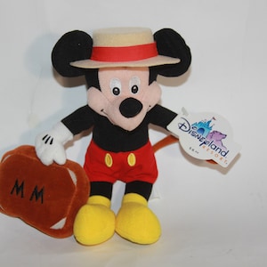 Puede incluir: Un juguete de peluche de Mickey Mouse que lleva un sombrero de paja con una banda roja. Lleva una maleta marr&oacute;n con las iniciales "MM" en ella. El juguete tiene una etiqueta de Disneyland Resort adjunta.