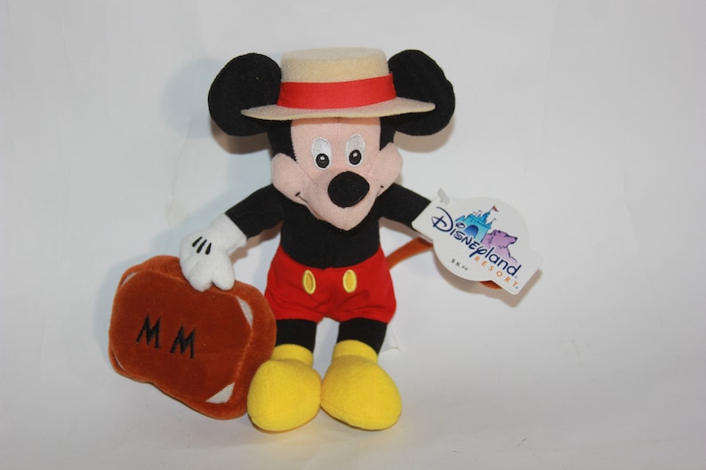 Puede incluir: Un juguete de peluche de Mickey Mouse que lleva un sombrero de paja con una banda roja. Lleva una maleta marr&oacute;n con las iniciales "MM" en ella. El juguete tiene una etiqueta de Disneyland Resort adjunta.