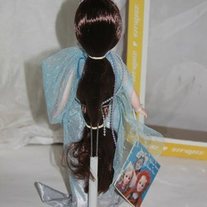 May include: A porcelain doll with long brown hair wearing a light blue gown with a silver trim. The doll is on a stand and has a tag that says "It's a Girl!" with a picture of two dolls.