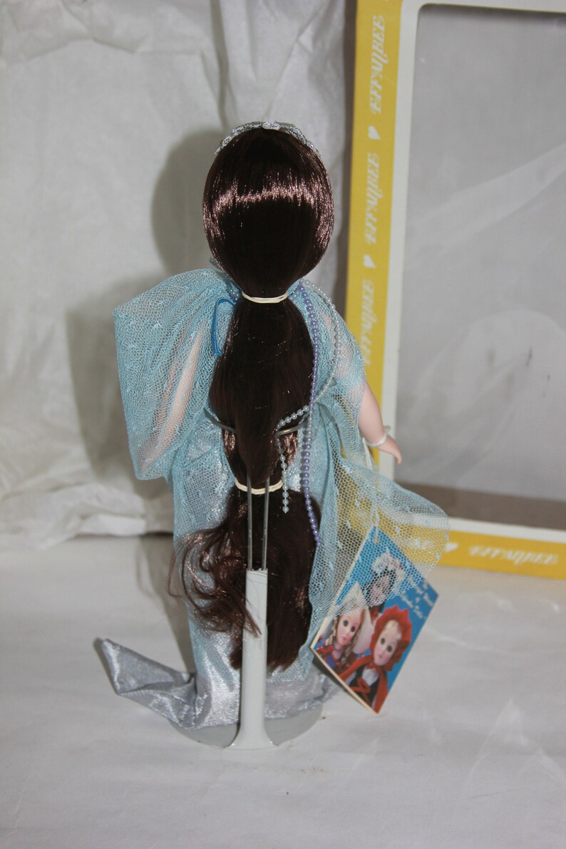May include: A porcelain doll with long brown hair wearing a light blue gown with a silver trim. The doll is on a stand and has a tag that says "It's a Girl!" with a picture of two dolls.