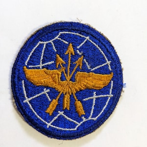 May include: A blue and gold embroidered patch with a winged eagle and arrows. The eagle is holding three arrows in its talons, and the arrows are pointing upwards. The patch is surrounded by a white circle.