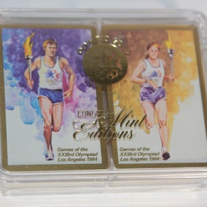 May include: Two playing cards in a clear plastic case. The cards feature a watercolor illustration of a runner carrying a torch. The cards are gold with the text "Games of the XXIIIrd Olympiad Los Angeles 1984" and "Congressional Mint Editions".