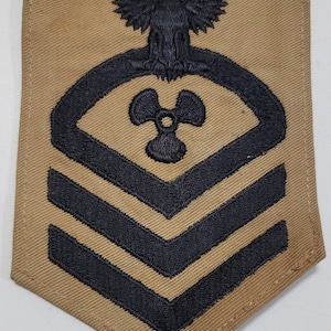 May include: A tan fabric patch with a black embroidered eagle, propeller, and three chevrons. The patch is shaped like a diamond.