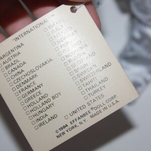 May include: A white tag with a list of countries printed in black. The tag reads "International" and lists countries including Argentina, Austria, Brazil, Canada, China, Czechoslovakia, Denmark, France, Germany, Greece, Holland, Holland Boy, Hungary, India, Ireland, Israel, Italy, Mexico, Norway, Peru, Poland, Romania, Russia, Scotland, Spain, Sweden, Switzerland, Tahiti, Thailand, Turkey, United States. The tag also includes the copyright information "&copy;1984 EFFANBEE&reg; DOLL CORP. NEW YORK, N.Y. MADE IN U.S.A."