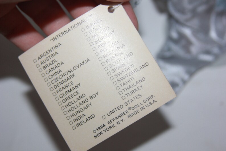 May include: A white tag with a list of countries printed in black. The tag reads "International" and lists countries including Argentina, Austria, Brazil, Canada, China, Czechoslovakia, Denmark, France, Germany, Greece, Holland, Holland Boy, Hungary, India, Ireland, Israel, Italy, Mexico, Norway, Peru, Poland, Romania, Russia, Scotland, Spain, Sweden, Switzerland, Tahiti, Thailand, Turkey, United States. The tag also includes the copyright information "&copy;1984 EFFANBEE&reg; DOLL CORP. NEW YORK, N.Y. MADE IN U.S.A."