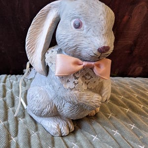 May include: A light gray ceramic bunny figurine with a pink bow tie and a white lace collar. The bunny has long ears and a detailed textured surface. The figurine is sitting on a green quilted surface.