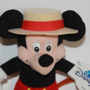 Puede incluir: Un juguete de peluche de Mickey Mouse que lleva un sombrero de color beige con una banda roja. El juguete tiene una nariz negra, ojos negros y una cara rosa. El juguete sostiene un palo marr&oacute;n en su mano derecha. El juguete tiene una etiqueta blanca pegada a su brazo izquierdo que dice "Disneyland Resort".
