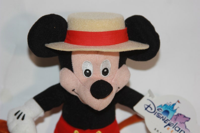 Puede incluir: Un juguete de peluche de Mickey Mouse que lleva un sombrero de color beige con una banda roja. El juguete tiene una nariz negra, ojos negros y una cara rosa. El juguete sostiene un palo marr&oacute;n en su mano derecha. El juguete tiene una etiqueta blanca pegada a su brazo izquierdo que dice "Disneyland Resort".