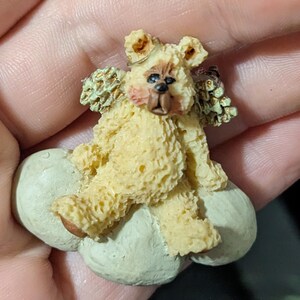 May include: A small, detailed figurine of a teddy bear angel. The bear is light yellow with a textured surface, sitting on a light green cloud. It has small, detailed wings and a sweet expression.