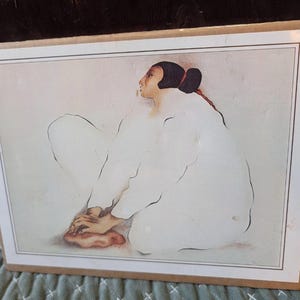 May include: A framed print featuring a seated figure in a white garment. The figure has dark hair pulled back, with a red accent. The artwork has a soft, muted color palette, with the figure's hands resting on their knees.
