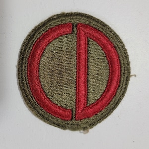 May include: A green and red military patch with a red "D" inside a red circle.