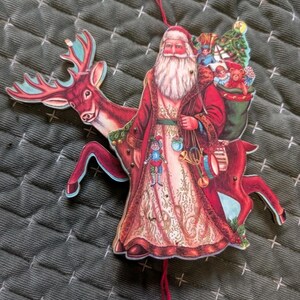 May include: A flat, die-cut hanging ornament featuring a vintage-style Santa Claus in a red coat with a white beard, standing beside a red reindeer. Santa carries a green sack filled with toys and holds a small drum and horn, with a red string pull.