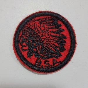 May include: A red and black embroidered patch with an image of an American Indian wearing a headdress. The patch has the letters "BSA" embroidered in the bottom center.