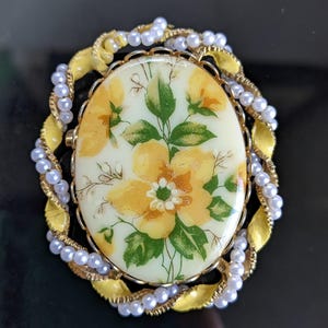 May include: Oval-shaped vintage brooch with a floral design. The center features a cream-colored background with yellow flowers and green leaves. The brooch is framed by a gold-colored metal border, a yellow leaf design, and a strand of small white pearls.