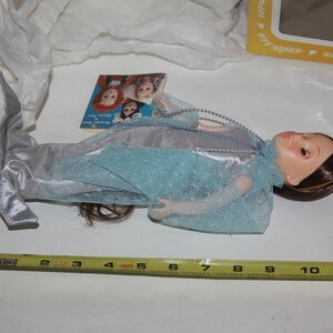 May include: A blue and silver mermaid doll with long brown hair and a crown. The doll is lying on a white surface with a measuring tape and a small book.