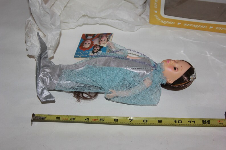May include: A blue and silver mermaid doll with long brown hair and a crown. The doll is lying on a white surface with a measuring tape and a small book.