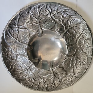 May include: A silver metal bowl with a decorative design of leaves. The bowl is round and has a smooth, shiny surface.