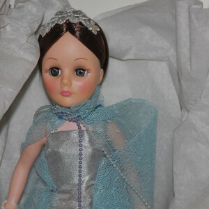 May include: A porcelain doll with brown hair, wearing a silver dress and a light blue lace shawl. The doll has a pearl necklace and a silver crown.