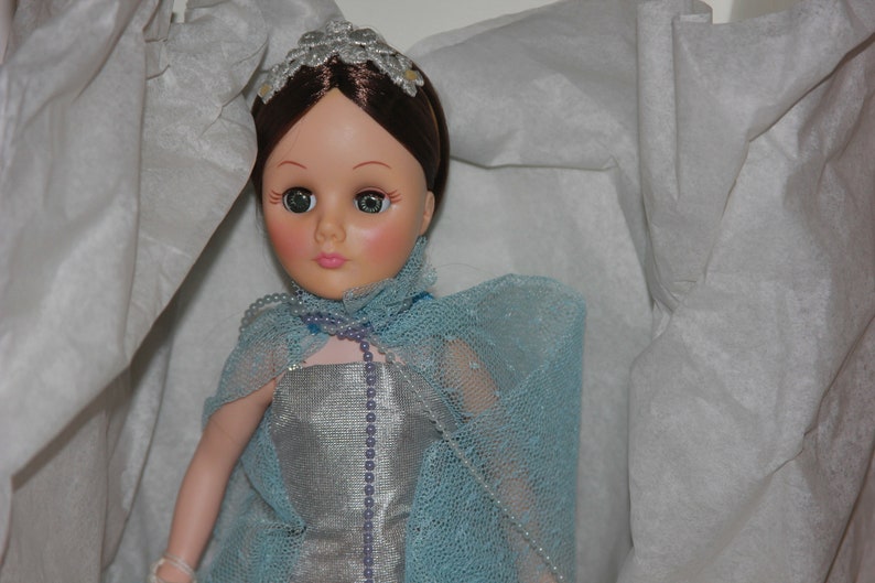 May include: A porcelain doll with brown hair, wearing a silver dress and a light blue lace shawl. The doll has a pearl necklace and a silver crown.