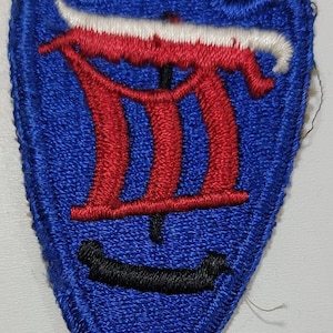 May include: Embroidered blue patch with a stylized design featuring red and white elements. The patch has a unique shape, with a blue arrowhead base and a white and red design at the top. A black shape is at the bottom.