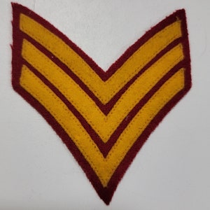 May include: A felt chevron patch with a burgundy border and three gold stripes. The patch is in a V-shape, commonly used to denote rank or service in military or other uniformed organizations.