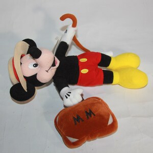 Puede incluir: Un juguete de peluche de Mickey Mouse con un sombrero marr&oacute;n y pantalones cortos rojos. Lleva una maleta marr&oacute;n con las iniciales "MM" bordadas. Tambi&eacute;n lleva un bast&oacute;n marr&oacute;n.