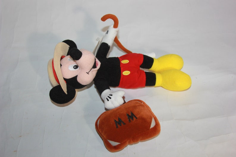 Puede incluir: Un juguete de peluche de Mickey Mouse con un sombrero marr&oacute;n y pantalones cortos rojos. Lleva una maleta marr&oacute;n con las iniciales "MM" bordadas. Tambi&eacute;n lleva un bast&oacute;n marr&oacute;n.