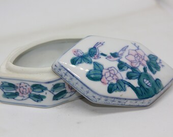 May include: A white porcelain trinket box with a hexagonal lid. The lid is decorated with a floral design featuring pink flowers, green leaves, and blue birds. The box has a similar floral design on the sides.