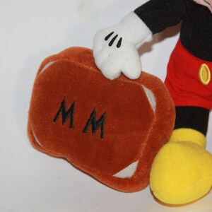 Puede incluir: Un juguete de peluche de Mickey Mouse sosteniendo una maleta marr&oacute;n con las iniciales "MM" bordadas. La maleta tiene ribetes blancos. Mickey lleva pantalones cortos rojos con botones amarillos, zapatos negros con suelas amarillas y guantes blancos.