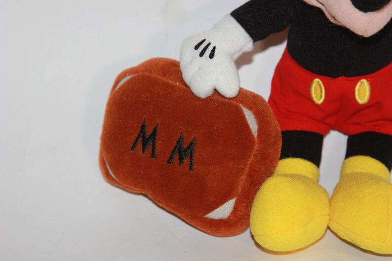 Puede incluir: Un juguete de peluche de Mickey Mouse sosteniendo una maleta marr&oacute;n con las iniciales "MM" bordadas. La maleta tiene ribetes blancos. Mickey lleva pantalones cortos rojos con botones amarillos, zapatos negros con suelas amarillas y guantes blancos.