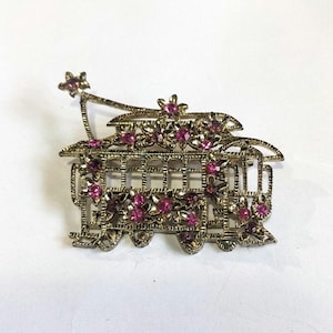 May include: A gold-tone brooch in the shape of a vintage trolley car, decorated with pink rhinestones. The trolley car has a star-shaped detail on top.