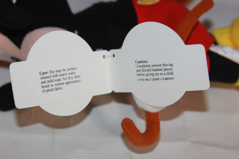 Puede incluir: Etiquetas blancas de cuidado y precauci&oacute;n adjuntas a un juguete de peluche de Mickey Mouse. La etiqueta de cuidado indica que se debe limpiar la superficie con agua tibia y jab&oacute;n suave, secar al aire y cepillar para restaurar la apariencia del tejido de felpa. La etiqueta de precauci&oacute;n indica que se debe quitar completamente la etiqueta y desechar las piezas de sujeci&oacute;n antes de dar el juguete a un ni&ntilde;o. La etiqueta tambi&eacute;n incluye el texto "THE WALT DISNEY COMPANY".