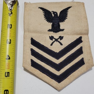 May include: A beige fabric patch with a black eagle, crossed axes, and three black chevrons. The patch is a military insignia.