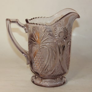 May include: A clear glass pitcher with a delicate, intricate pattern. The pitcher is a pale lavender color and has a footed base.