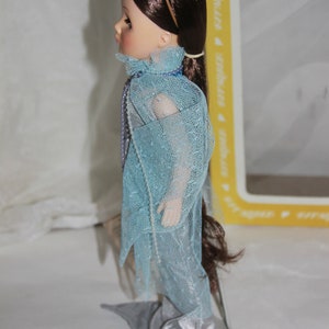 May include: A porcelain doll with long brown hair wearing a light blue dress with a silver trim. The doll is wearing a silver headband with a flower design.
