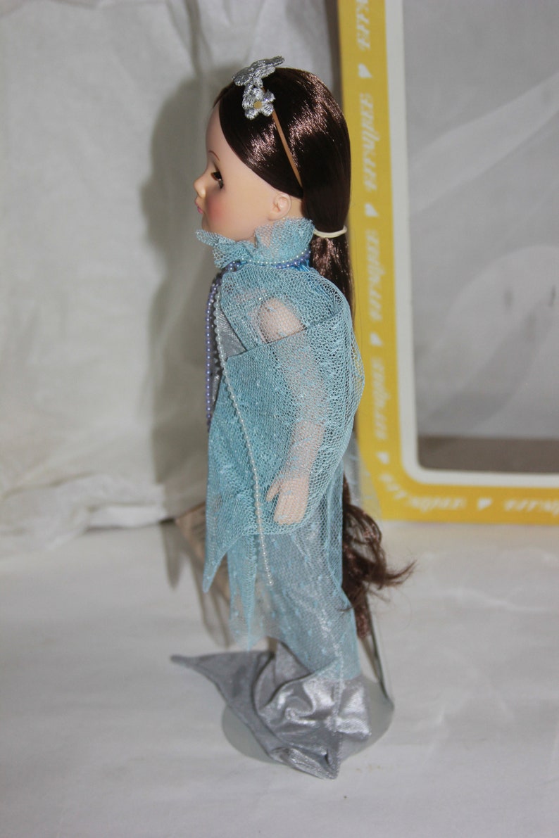 May include: A porcelain doll with long brown hair wearing a light blue dress with a silver trim. The doll is wearing a silver headband with a flower design.