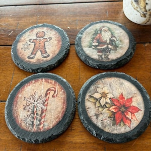 May include: Four round slate coasters with Christmas-themed designs. One features a gingerbread man, another Santa Claus, a candy cane, and a poinsettia. Each coaster has a dark, textured border and a vintage-style print.