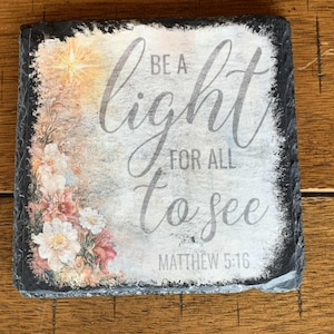 Puede incluir: Posavasos cuadrado de pizarra con el texto "Be a light for all to see" y "Matthew 5:16". El posavasos presenta un diseño floral con flores blancas, rosas y melocotón, y una estrella.