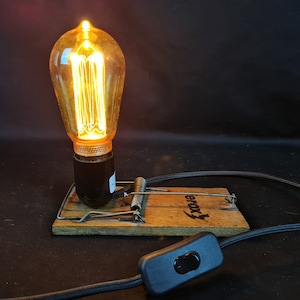 Op de afbeelding: Een vintage-stijl gloeilamp gemonteerd op een houten muizenvalbasis met een zwart snoer en een aan/uit-schakelaar. De gloeilamp is aan en heeft een warme gloed.