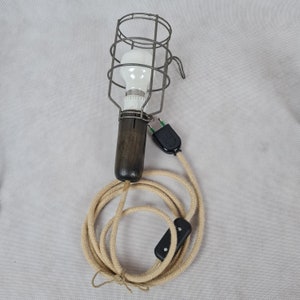 Peut inclure: Une lampe de travail vintage avec une ampoule en cage et une poignée en bois brun. La lampe est reliée à un long cordon beige avec un interrupteur noir.