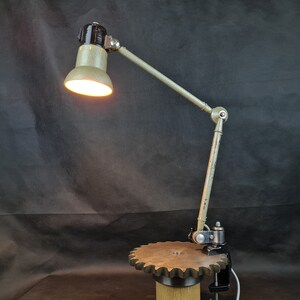 May include: A vintage industrial style desk lamp with a gray metal shade and a black metal base. The lamp is mounted on a metal arm with a gear-shaped clamp.