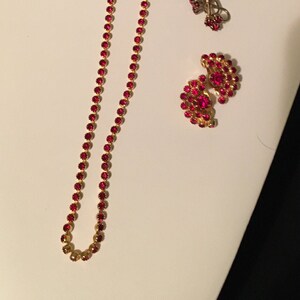 May include: A set of red rhinestone jewelry featuring a necklace, a pair of earrings, and a brooch. The necklace is a simple chain of red rhinestones. The earrings are a pair of red rhinestone flower shapes. The brooch is a heart shape with red rhinestones.
