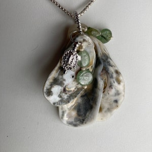 May include: A silver necklace with a pendant made from a natural oyster shell. The shell is white and grey with a silver turtle charm, green beads, and a silver chain. The necklace is a unique piece of jewellery.