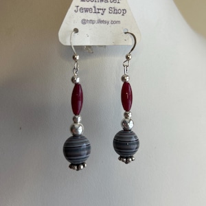 May include: A pair of dangle earrings with silver-tone hooks. Each earring features a dark red oval bead, a silver bead, a striped gray and black round bead, and a silver-tone decorative base. The earrings are displayed on a white surface.