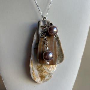 Pode incluir: Um colar de corrente prateada com um pingente feito de três conchas de ostra sobrepostas. As conchas são marrons, brancas e douradas com um brilho perolado. O pingente é acentuado por duas pequenas pérolas roxas escuras e uma conta de cristal.