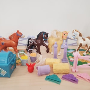 Könnte beinhalten: Ein Set aus Plastik-Spielzeugpferden und Zubehör, darunter ein braunes Pferd, ein schwarz-weißes Pferd, ein weißes Pferd, eine blaue Truhe, ein lila Eimer und ein gelber Eimer. Die Pferde stehen auf einem rosa-weißen Plastikzaun.