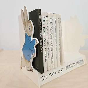 May include: A white wooden bookend featuring a Peter Rabbit figure in a blue jacket. The bookend holds a collection of Beatrix Potter books, including "The Tale of Two Bad Mice" and "The Tale of Peter Rabbit". The base reads "The World of Beatrix Potter".