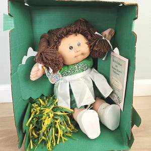 May include: A Cabbage Patch Kids doll with brown yarn hair in pigtails, wearing a green and white cheerleader outfit. The doll is inside a green cardboard box with a yellow and green pom-pom, a birth certificate, and adoption papers.