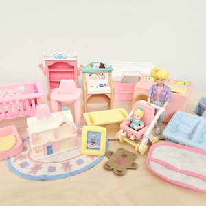 May include: A collection of plastic dollhouse furniture and accessories including a pink crib, a high chair, a small house, a yellow table, a stroller with a baby doll, a larger doll, a blue bathtub, a rug, and a small picture frame.